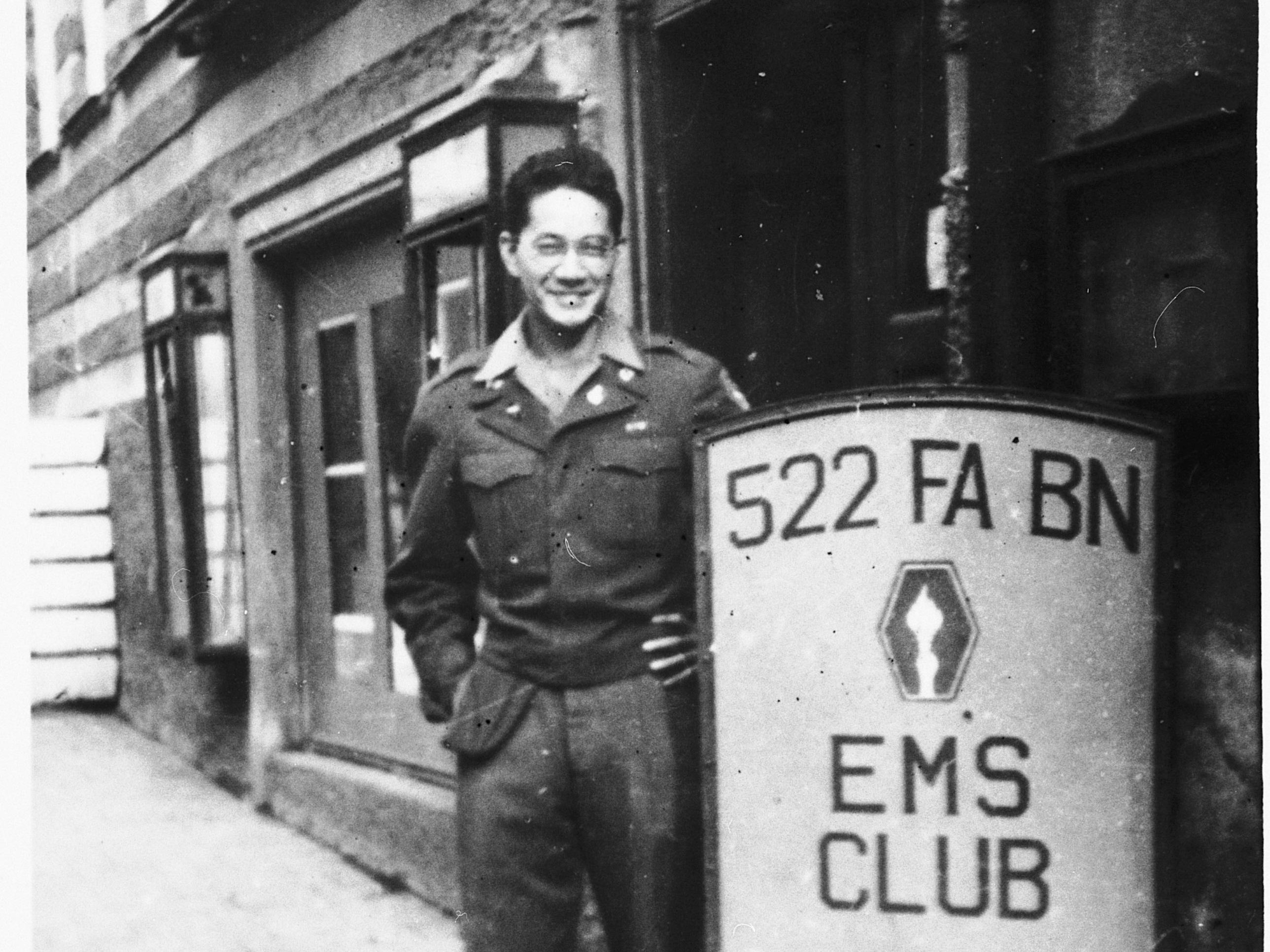 Clarence Matsumura, ein japanisch-amerikanischer Soldat im 522nd Field Artillery Battalion in Deutschland, posiert neben einem Schild des 522 EMS Club. Matsumura war der Soldat, der den jüdischen Überlebenden Solly Ganor von einem Todesmarsch in Waakirchen befreite.